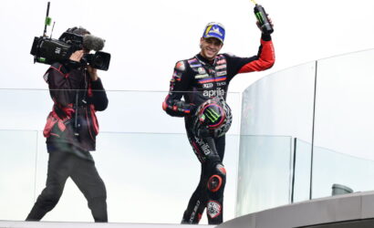 MotoGP rider in black/red Aprilia suit on a glass podium, waving a Monster energy bottle as a cameraman films him.