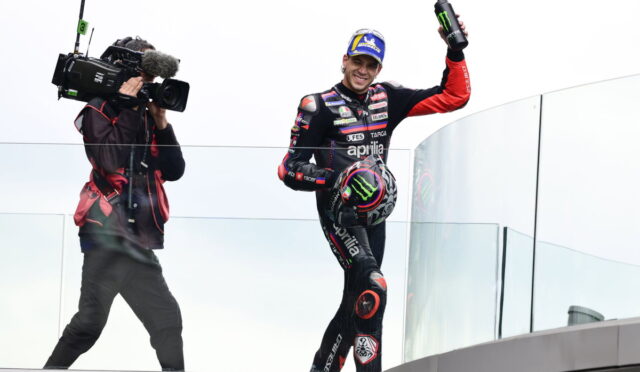 MotoGP rider in black/red Aprilia suit on a glass podium, waving a Monster energy bottle as a cameraman films him.