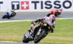 Moto racer in pink–black–yellow Monster livery leans into a turn on a race track; blurred riders in background.