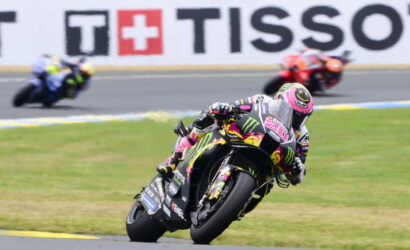 Moto racer in pink–black–yellow Monster livery leans into a turn on a race track; blurred riders in background.