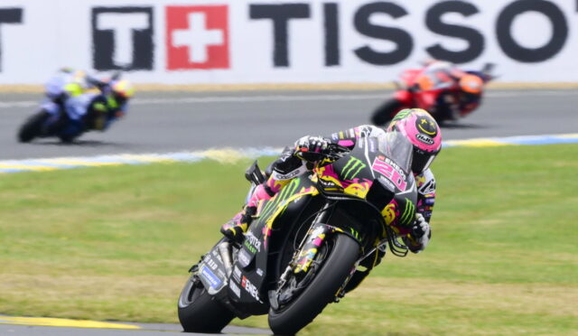 Moto racer in pink–black–yellow Monster livery leans into a turn on a race track; blurred riders in background.