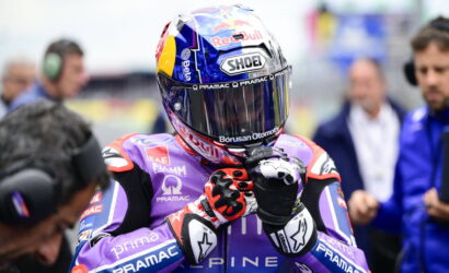 MotoGP rider in a purple Pramac racing suit and full-face helmet with Red Bull branding, standing in the pit area with gloved hands together in front of the chest.