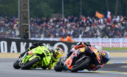 Two motorcycle racers lean into a sharp turn on a racetrack, neon-green and orange bikes in close pursuit while a blurred crowd watches from the stands behind them.