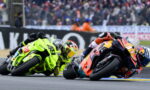 Two motorcycle racers lean into a tight turn on a race track, neon-green and orange-red bikes close together with a cheering crowd in the background.