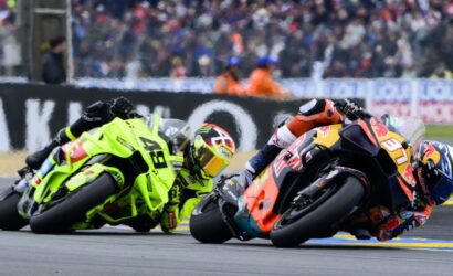 Two motorcycle racers lean into a tight turn on a race track, neon-green and orange-red bikes close together with a cheering crowd in the background.
