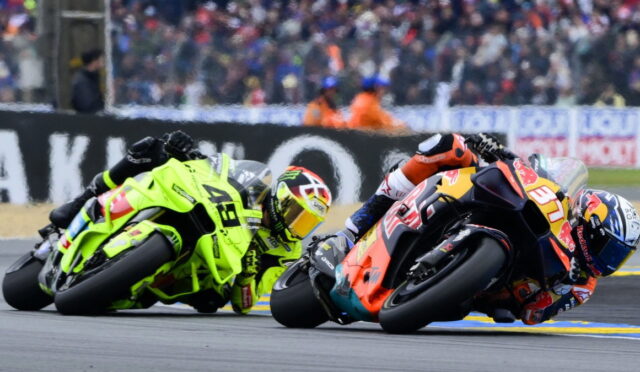 Two motorcycle racers lean into a tight turn on a race track, neon-green and orange-red bikes close together with a cheering crowd in the background.