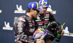Two motorcycle racers in racing suits share a smile, one holding a Red Bull helmet in front of a Michelin backdrop