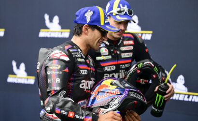 Two motorcycle racers in racing suits share a smile, one holding a Red Bull helmet in front of a Michelin backdrop