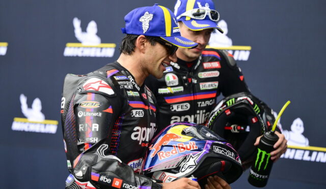 Two motorcycle racers in racing suits share a smile, one holding a Red Bull helmet in front of a Michelin backdrop
