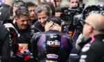 Motorsport team members comfort a rider in an Aprilia Racing suit as cameras surround them in the paddock scene.
