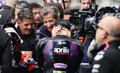 Motorsport team members comfort a rider in an Aprilia Racing suit as cameras surround them in the paddock scene.