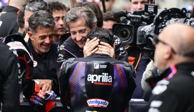 Motorsport team members comfort a rider in an Aprilia Racing suit as cameras surround them in the paddock scene.