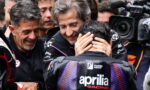 Rider in Aprilia Racing gear is consoled by two team members while cameras film the moment.