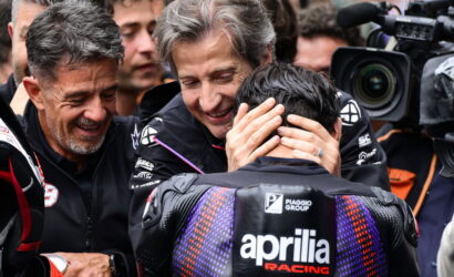 Rider in Aprilia Racing gear is consoled by two team members while cameras film the moment.