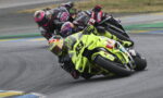 Front rider on a bright yellow race bike leans into a turn as a pack follows closely on a curving track.
