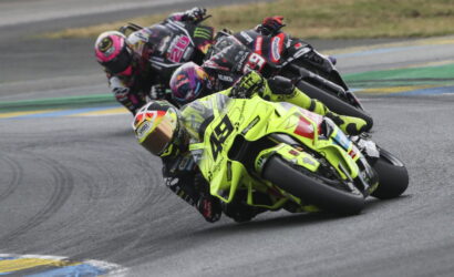 Front rider on a bright yellow race bike leans into a turn as a pack follows closely on a curving track.