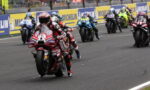 Motorcycle racers lean into a turn on a racing track, red Ducati leading the pack with Michelin barriers in the background