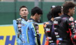 Motorsport riders in blue and black racing suits stand together on a track, facing different directions, with sponsor patches visible on their outfits.