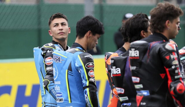 Motorsport riders in blue and black racing suits stand together on a track, facing different directions, with sponsor patches visible on their outfits.