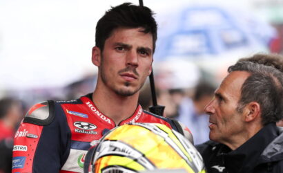 Young motorbike racer in red-and-blue suit listens to a man while holding a bright yellow helmet with black accents under a canopy.
