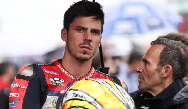 Young motorbike racer in red-and-blue suit listens to a man while holding a bright yellow helmet with black accents under a canopy.