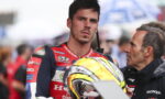 MotoGP rider in a red Honda racing suit holding his helmet, listening to a man in a jacket beside him.