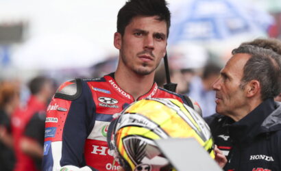 MotoGP rider in a red Honda racing suit holding his helmet, listening to a man in a jacket beside him.