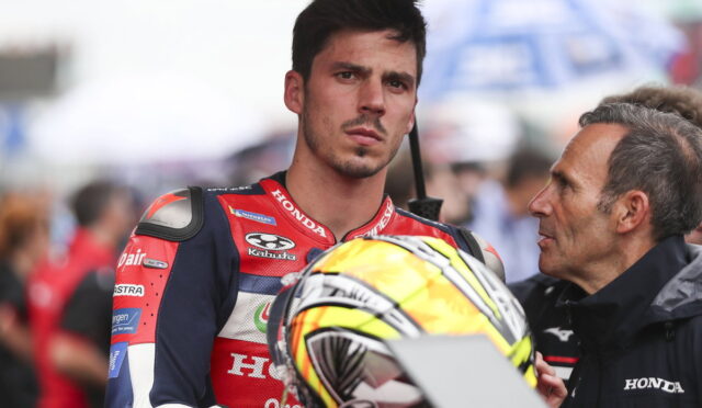 MotoGP rider in a red Honda racing suit holding his helmet, listening to a man in a jacket beside him.