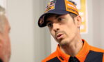 Man in KTM racing orange gear and cap leaning in as he talks to another person.