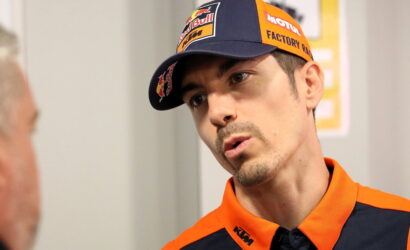 Man in KTM racing orange gear and cap leaning in as he talks to another person.