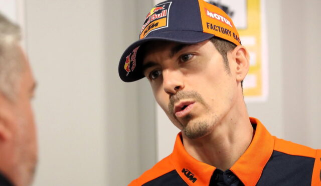 Man in KTM racing orange gear and cap leaning in as he talks to another person.