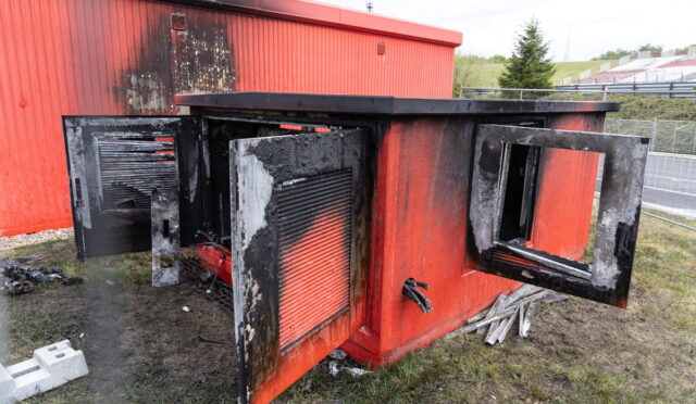 Red outdoor metal cabinet heavily burned, with doors open and charred interior panels visible.