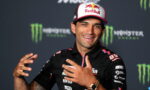 Smiling racing athlete in a Red Bull cap and black sponsor polo at a press event, with Monster Energy backdrop behind him.