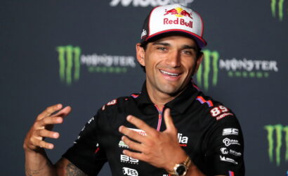 Smiling racing athlete in a Red Bull cap and black sponsor polo at a press event, with Monster Energy backdrop behind him.