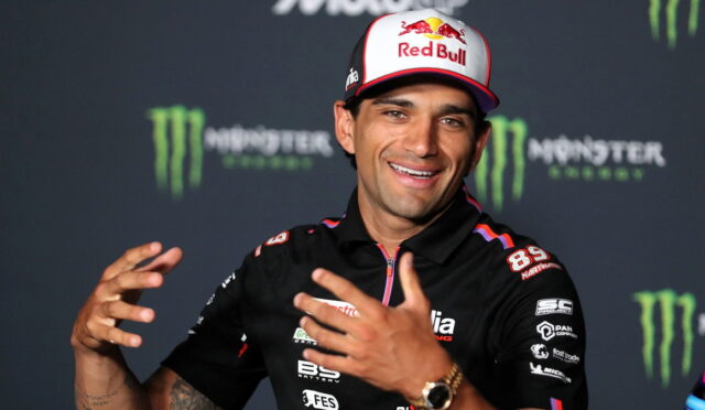Smiling racing athlete in a Red Bull cap and black sponsor polo at a press event, with Monster Energy backdrop behind him.