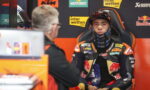 Rider in full racing gear sits for an interview in the pit area, sponsor banners in the background left and center.