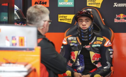 Rider in full racing gear sits for an interview in the pit area, sponsor banners in the background left and center.