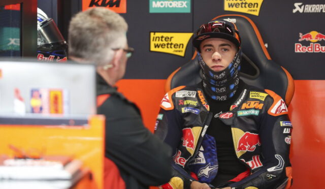 Rider in full racing gear sits for an interview in the pit area, sponsor banners in the background left and center.