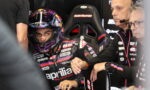 MotoGP rider in full race gear and helmet sits in a garage, ready for a session, APRILIA Racing branding visible on suit and gloves with team members nearby.