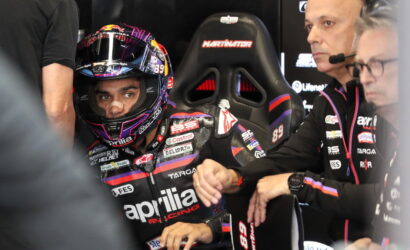 MotoGP rider in full race gear and helmet sits in a garage, ready for a session, APRILIA Racing branding visible on suit and gloves with team members nearby.