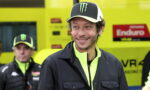 Smiling man in a black jacket and neon yellow zip-up, wearing a black cap with a green Monster logo, in a bright yellow racing pit area.
