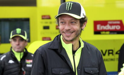 Smiling man in a black jacket and neon yellow zip-up, wearing a black cap with a green Monster logo, in a bright yellow racing pit area.