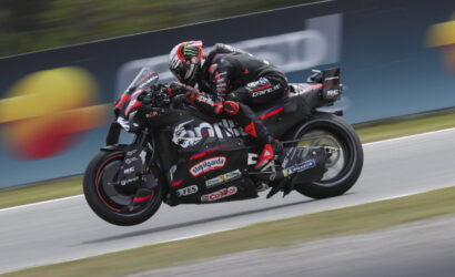 Rider in black racing suit on a black and red race bike with sponsor stickers, speeding on a track.