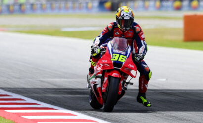 Moto racer in red-white-blue leathers leans into a left-hand turn on a track; bike number 36 visible on the red bike with sponsor logos.