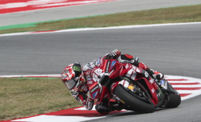 Motorcycle racer leaning into a left-hand turn on a race track, red-and-white livery with sponsor logos visible beneath the rider's helmet.