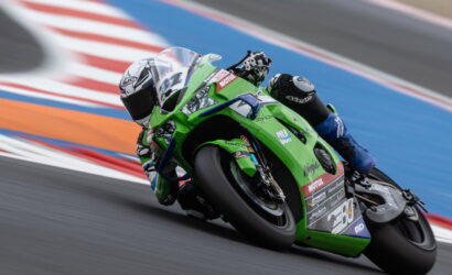 Rider on a bright green race bike leans into a sharp turn on a racetrack, blue run-off and red/white curbs in the background.