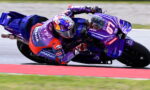 Moto GP rider in purple Pramac livery leans into a corner on a blue-purple Yamaha bike, helmet with colorful sponsor decals, mid-race.