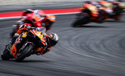 MotoGP-style race: rider in orange suit leaning into a sharp turn, number 37 on the bike, with blurred pursuers in the background on a gray track.