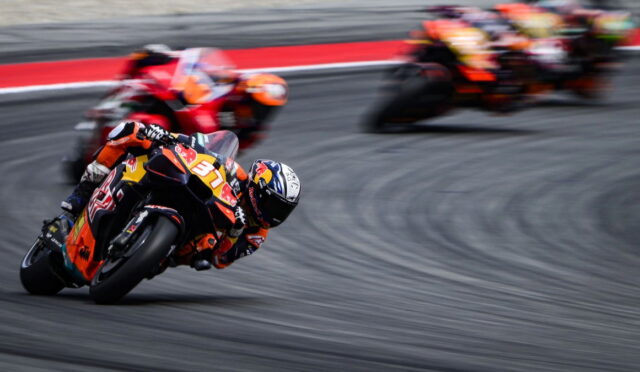 MotoGP-style race: rider in orange suit leaning into a sharp turn, number 37 on the bike, with blurred pursuers in the background on a gray track.