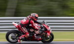 MotoGP rider in red livery leaning into a high-speed turn on a race track, blurred background behind metal barriers.